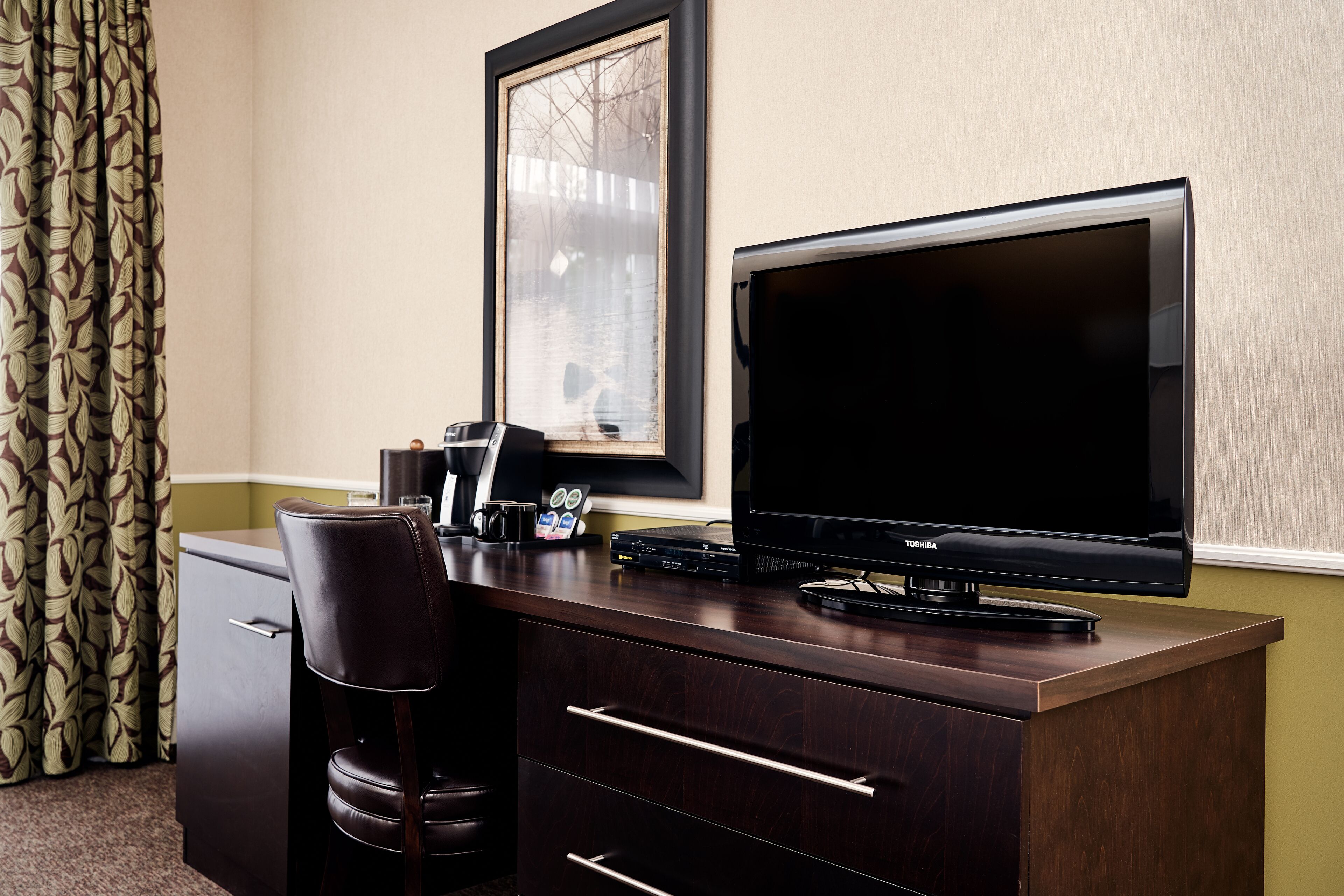 Una habitación de hotel con un escritorio de madera oscura, un televisor negro, un cuadro en la pared, una cafetera pequeña y una cortina estampada.