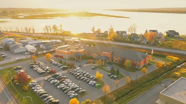 A large parking lot with multiple rows of cars, a building with a flat roof, and a body of water in the background. - RiverRock Inn & Conference Centre (Clarence-Rockland)