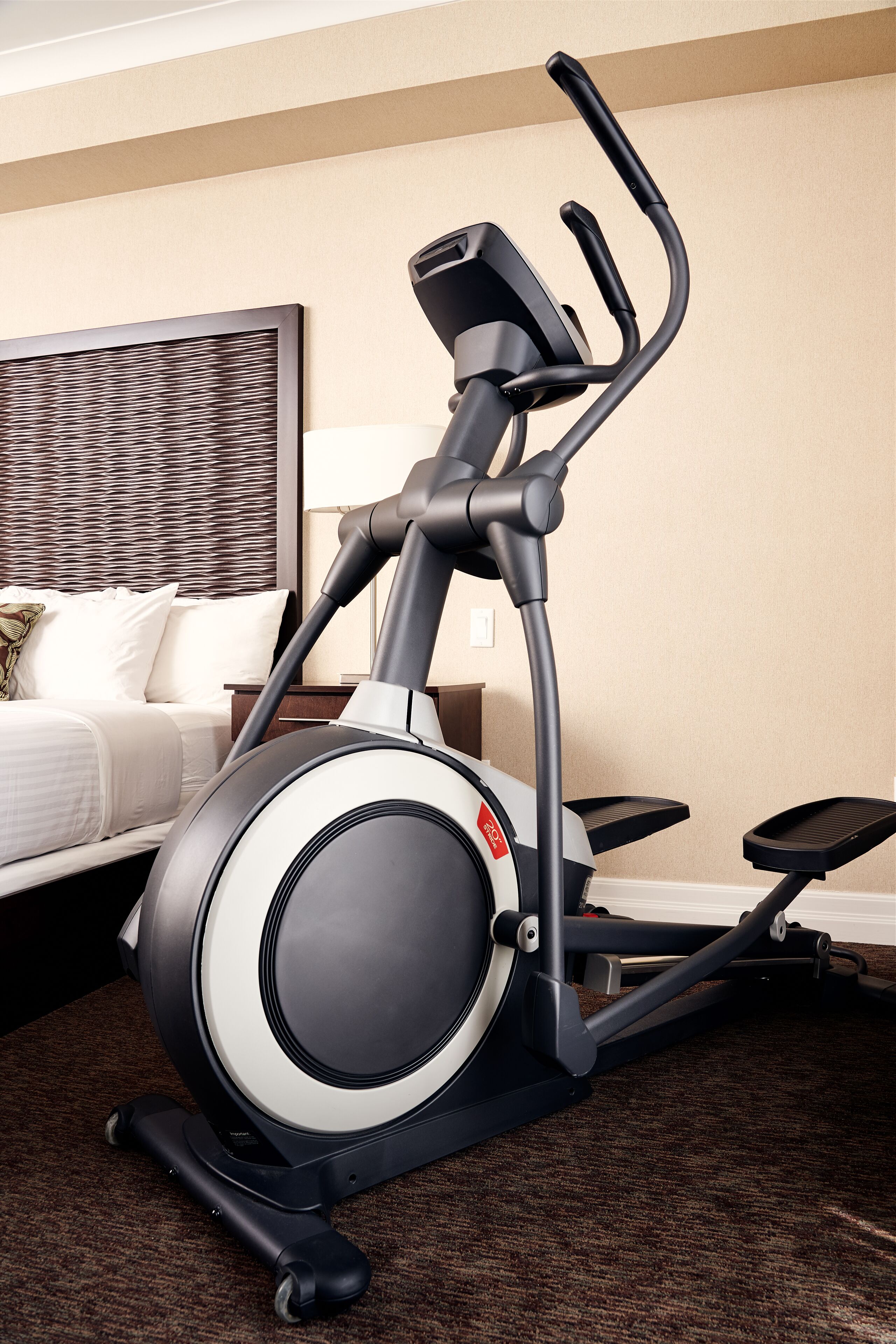 An exercise machine in a hotel room with a bed, a nightstand, and a wall-mounted lamp.