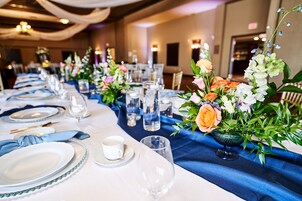 Uma mesa de banquete posta com pratos brancos, guardanapos azuis e copos de vidro transparente, decorada com arranjos florais.