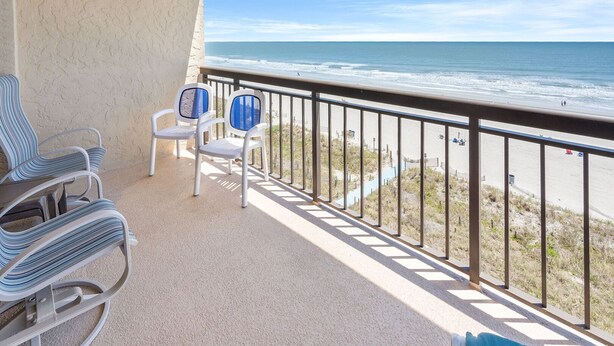 Ein Balkon mit zwei Stühlen und einem Tisch, der auf einen Strand mit freiem Blick auf das Meer und einen sandigen Uferbereich blickt.