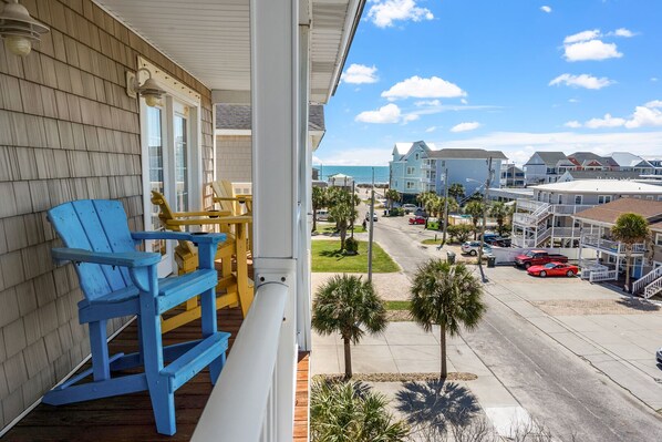 Een balkon met blauwe en gele Adirondack stoelen, uitzicht op een straat met palmbomen en een strand op de achtergrond.