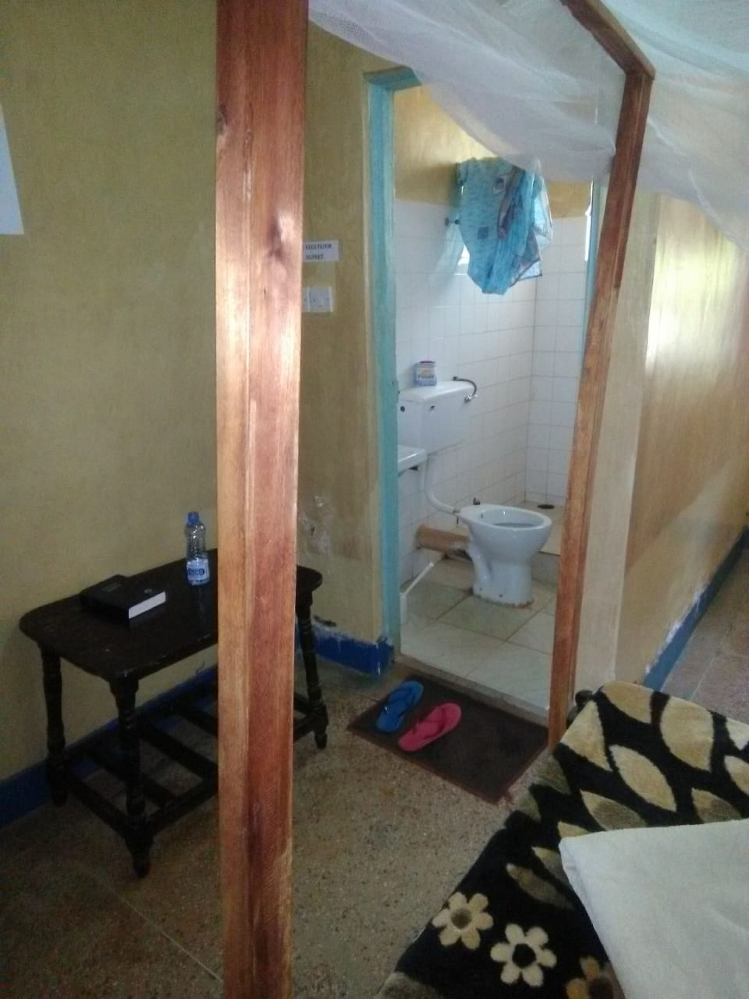 Un baño pequeño y funcional con inodoro, lavamanos y espejo. Hay una mesita de madera con una botella de agua y un libro encima. El suelo tiene una alfombra estampada, y hay una puerta que conduce a otra habitación.