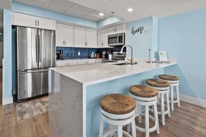 A modern kitchen with a stainless steel refrigerator, a microwave, and a large island with bar stools. - Discover Comfort & Convenience (North Myrtle Beach)