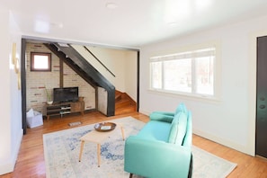 A living room with a teal sofa, a TV on a wooden stand, a staircase, and a window with blinds.