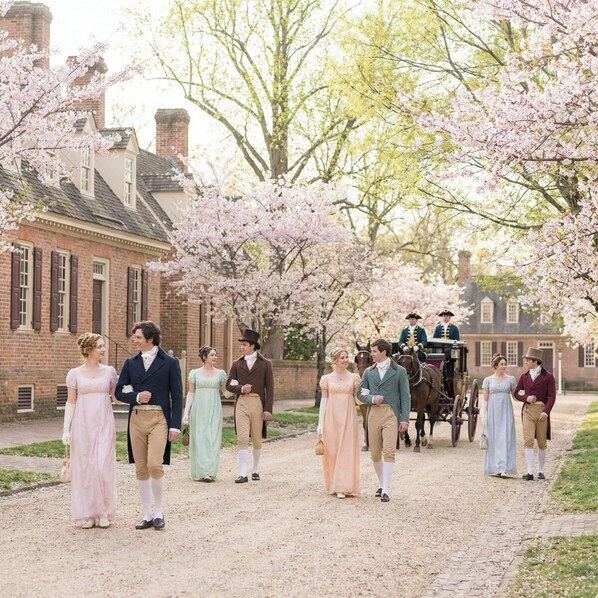Un groupe de personnes vêtues de costumes d’époque se promène le long d’un sentier bordé de cerisiers en fleurs, et une calèche tirée par des chevaux passe à proximité.
