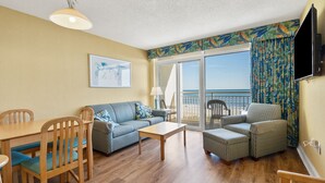 A room with a wooden dining table, a sofa, and two armchairs. There is a large window with a view of the beach.