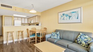 A compact living space with a kitchenette, a dining area with wooden chairs, a sofa with patterned cushions, and a framed artwork on the wall.