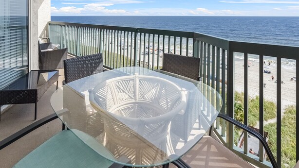 Ein Balkon mit Glastisch und Stühlen, der Blick auf einen Strand mit Menschen und einen klaren Himmel bietet.