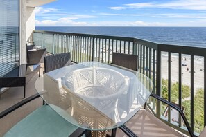 En balkon med et glasbord og stole, der kigger ud over en strand med mennesker og en klar himmel.