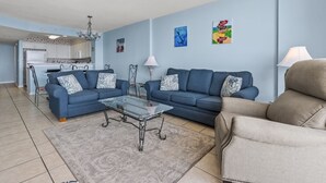 Ein Wohnzimmer mit zwei Sofas, einem Couchtisch und einem beigefarbenen Sessel. Im Hintergrund ist eine Küche mit weißen Schränken und einer Kronleuchter zu sehen.