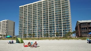 Un hôtel de grande hauteur doté de nombreux balcons, une plage de sable en premier plan et un ciel bleu dégagé.