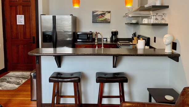 A modern kitchen with a stainless steel refrigerator, a microwave, and a coffee maker. There are two bar stools and a small table. - Beautiful center of Kalispell apartment (Kalispell)