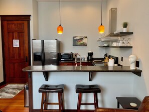 A modern kitchen with a stainless steel refrigerator, a microwave, and a coffee maker. There are two bar stools and a small table. - Beautiful center of Kalispell apartment (Kalispell)