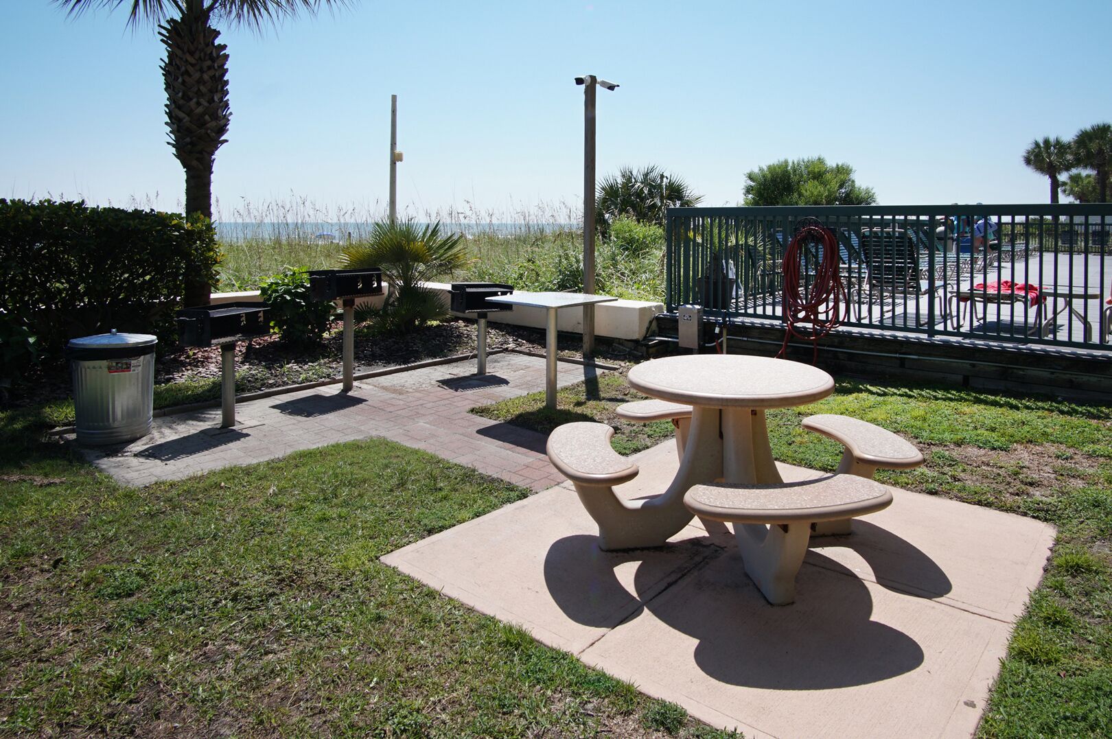 An outdoor patio area with a round table and benches, surrounded by a fence and palm trees.