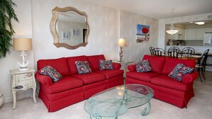 Un salón con dos sofás rojos, una mesa de centro de cristal, una lámpara blanca, un espejo decorativo y un cuadro en la pared.