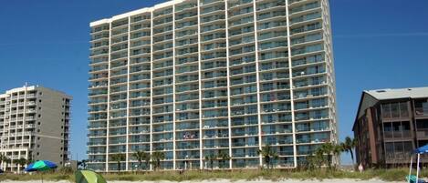 A high-rise hotel with multiple balconies, a sandy beach in the foreground, and a clear blue sky.