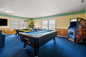 A pool table in a room with windows, a television, and arcade games.