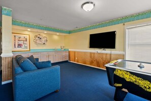 A room with a blue sofa, a flat-screen TV, a foosball table, and a kitchen area with wooden cabinets and a countertop. - Oceanfront duplex steps from beach w/shared pool, game room, kids' pool, grill (North Myrtle Beach)