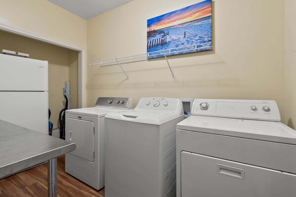 A laundry room with three white washing machines, a drying rack, and a painting of a beach scene on the wall.