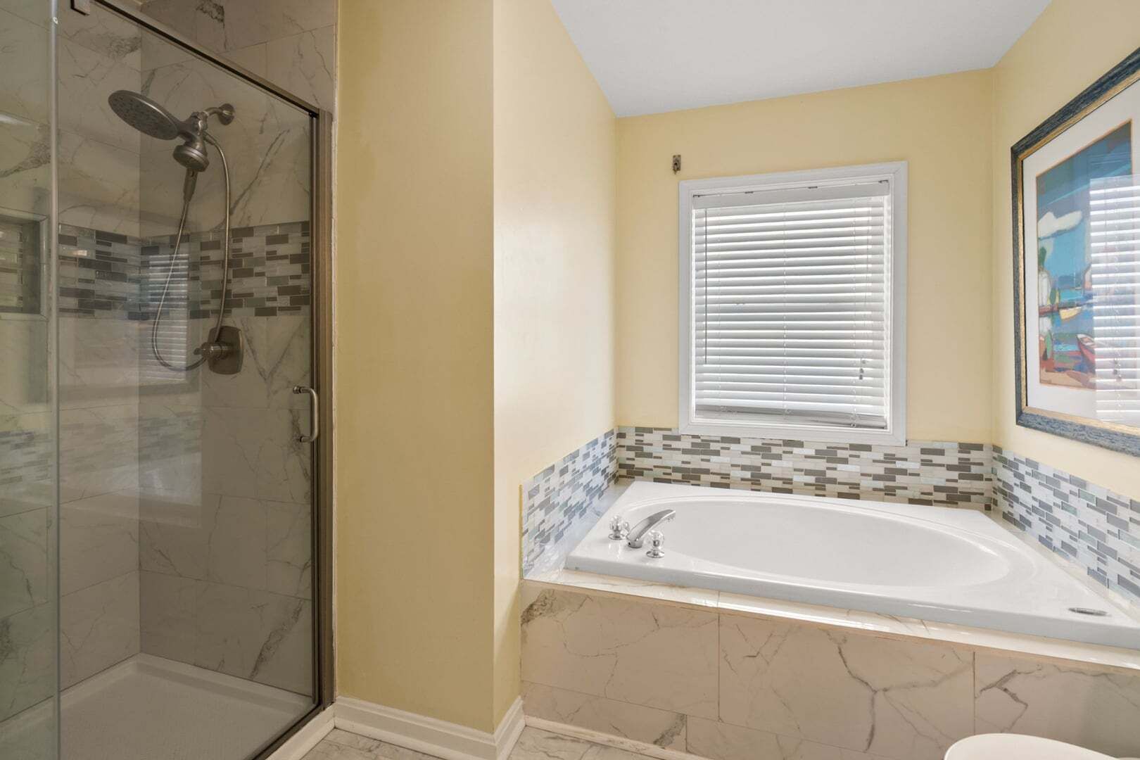 A bathroom with a glass shower enclosure, a bathtub with a mosaic tile surround, and a window with blinds.