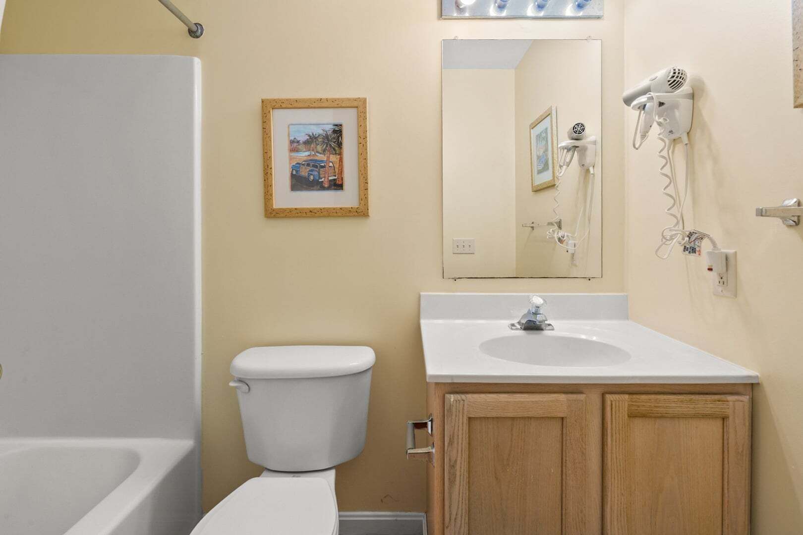 Une salle de bain avec des toilettes blanches, un lavabo surmonté d’un miroir et un sèche-cheveux fixé au mur.