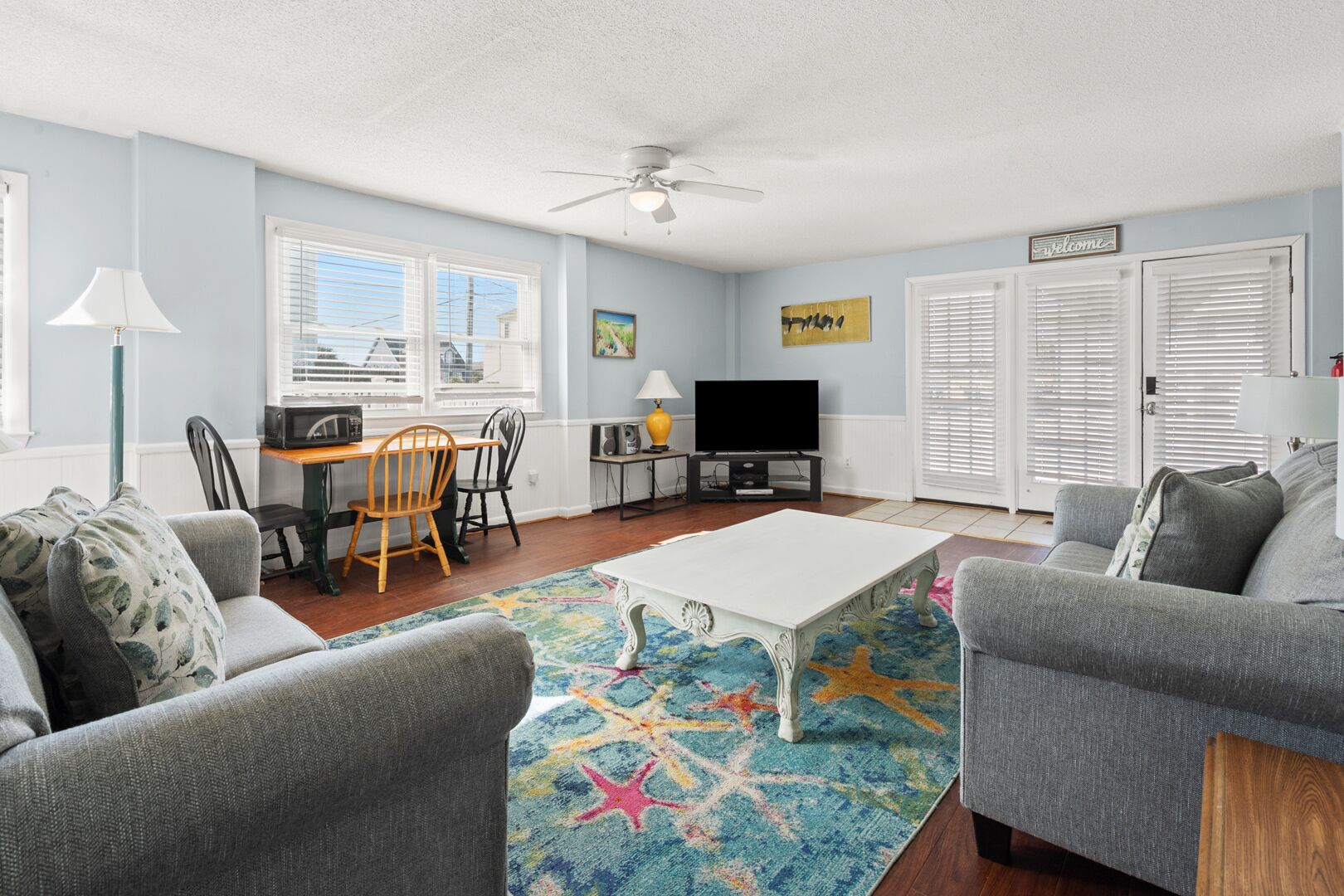 A living room with a white coffee table, grey sofas, a dining table with chairs, a TV, and a ceiling fan.