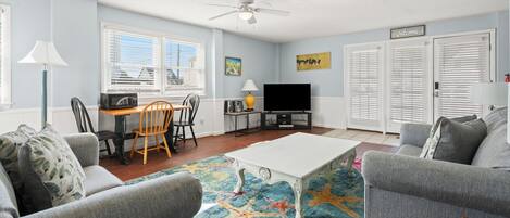 A living room with a white coffee table, grey sofas, a dining table with chairs, a TV, and a ceiling fan.