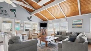 A spacious living room with a high ceiling, wooden beams, and a large window. There are two grey sofas, a coffee table, and a bookshelf. - Beautiful décor & large private pool! Spacious family home includes decks & huge yard in N. Myrtle. (North Myrtle Beach)