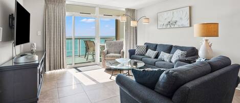 Un salon moderne avec un canapé, une table basse et un téléviseur posé sur un meuble. Il y a un balcon avec vue sur l’océan.