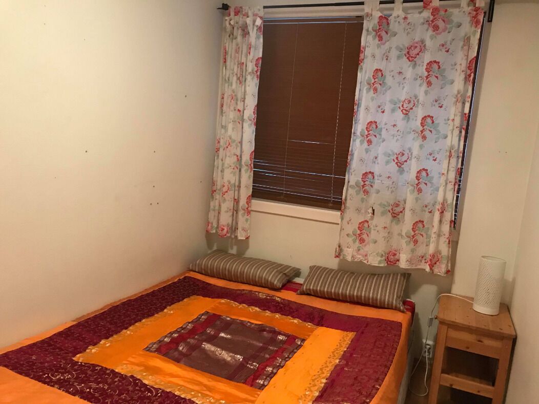 Une chambre à coucher avec un lit, des rideaux à motifs floraux, une fenêtre avec des stores et une table de chevet avec une lampe.