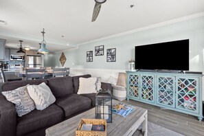 A modern living room with a grey sofa, a coffee table with a basket, a TV on a cabinet, and a dining area in the background. - Updated condo with amazing oceanfront view. Pool, sundeck, balconies perfect for couples or families (North Myrtle Beach)