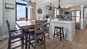 A modern kitchen with a large island, stainless steel appliances, and a dining area with dark chairs and a wooden table. - Updated condo with amazing oceanfront view. Pool, sundeck, balconies perfect for couples or families (North Myrtle Beach)