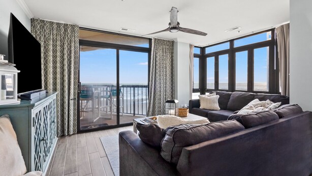A spacious living room with a large window, a ceiling fan, and a balcony with a view of the beach. - Updated condo with amazing oceanfront view. Pool, sundeck, balconies perfect for couples or families (North Myrtle Beach)
