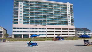 A large hotel with multiple floors and balconies, a parking garage, and beachside amenities including umbrellas and beach chairs.