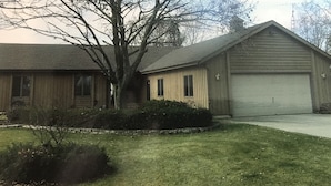 Uma casa de dois andares com garagem separada, uma grande árvore na frente e um gramado bem cuidado.