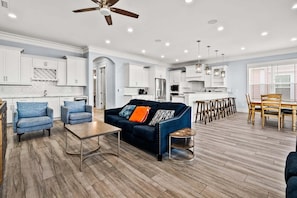 A modern kitchen with white cabinets, a dining area with wooden chairs, and a living room with a blue sofa and coffee table.