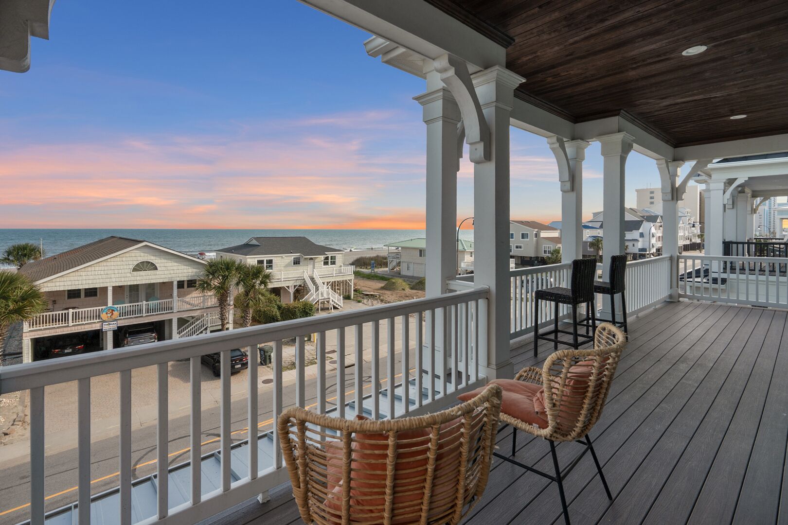 A covered outdoor area with a view of the beach, featuring a wooden deck, wicker chairs, and a table.