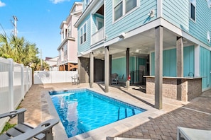 A swimming pool with a patio area, a covered outdoor seating space, and a two-story building with a light blue exterior.
