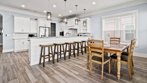 A modern kitchen with white cabinets, a central island, wooden stools, and a dining table with chairs. - Luxe boulevard home w/private pool, elevator, outdoor kitchen-purely wonderful! (North Myrtle Beach)