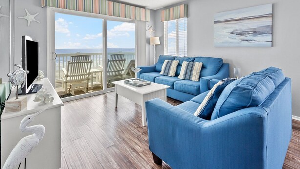 A living room with a blue sofa, a white coffee table, and a view of the beach through large windows.