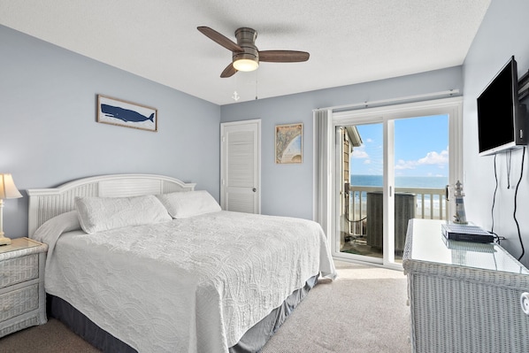 A bedroom with a bed, a ceiling fan, a desk with a computer, and a view of the ocean through a sliding door.