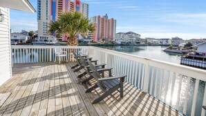 Une terrasse en bois avec des fauteuils Adirondack, un palmier et une vue sur une marina, avec des bâtiments en arrière-plan.
