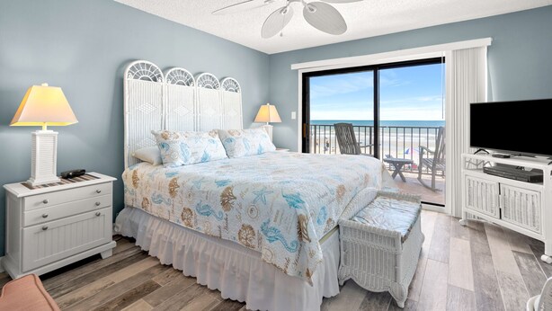 Een slaapkamer met een bed, een televisie, een balkon met uitzicht op het strand en een plafondventilator.