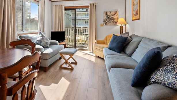 A living room with a wooden dining table, two sofas, a TV, and a balcony with a view. - Boulevard condo, steps from the beach, w/pool & sundeck, intimate setting for couples & families (North Myrtle Beach)