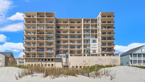 Een hotel met meerdere verdiepingen en balkons, gelegen aan een zandstrand met heldere lucht.