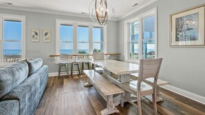 Een ruime woonkamer met een groot raam met uitzicht op de oceaan, een houten eettafel met banken en een grijze bank.