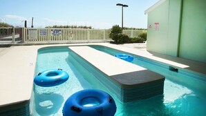 Une piscine avec deux bouées gonflables bleues, un poste de surveillance pour les sauveteurs et une zone délimitée par une clôture où sont affichées les règles de sécurité de la piscine.