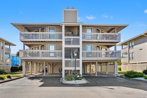 A multi-story residential building with balconies, a central entrance, and a parking area. - Cozy Boulevard condo--across street from ocean w/pool, sundeck, grill--waiting for your family! (North Myrtle Beach)
