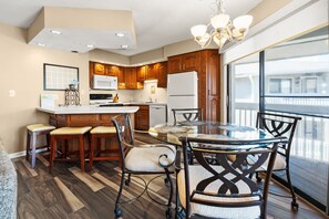 A modern kitchen with wooden cabinets, a central island, and a dining area with a glass table and chairs. - Cozy Boulevard condo--across street from ocean w/pool, sundeck, grill--waiting for your family! (North Myrtle Beach)